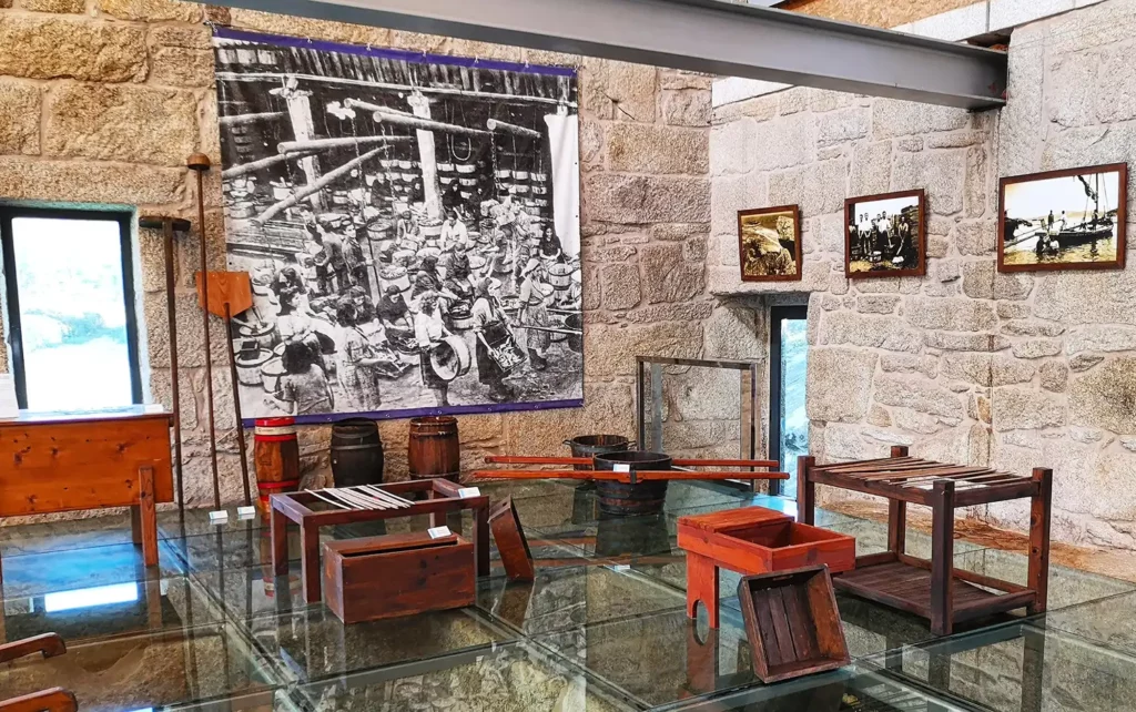 Sala de museo con pared de piedra, fotos enmarcadas, utensilios de madera y una gran imagen en blanco y negro de trabajadores.