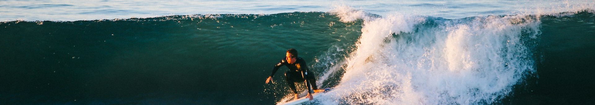 Surfista en traje de neopreno sobre una ola grande, con espuma blanca y fondo azul del mar bajo un cielo claro.